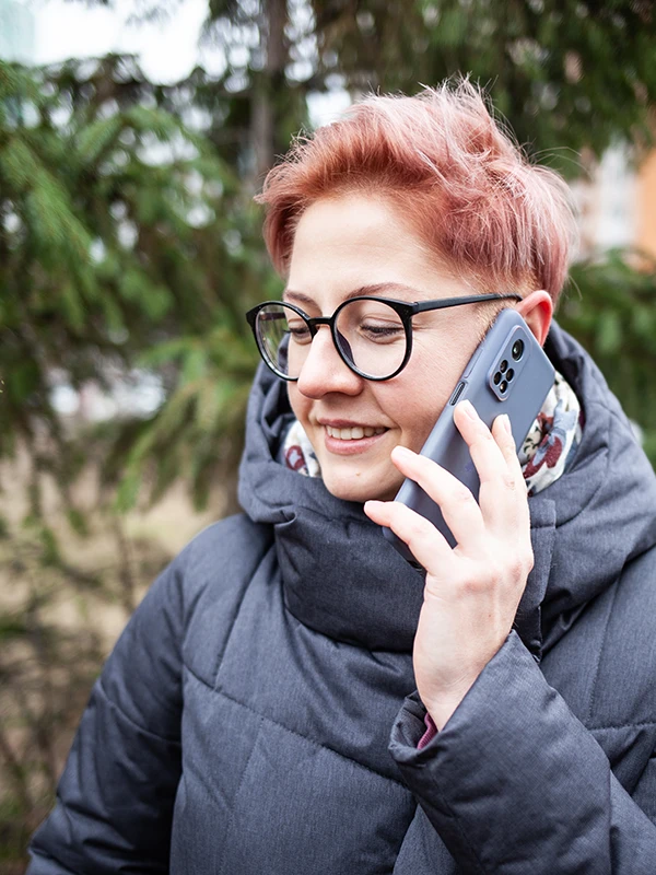 Naine lühikese roosa soengu ja prillidega räägib väljas nutitelefoniga, rohelised puud taustal