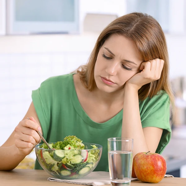 Naine istub laua ääres salatiga, pea toetatud käele, väsinud või tülpinud ilme