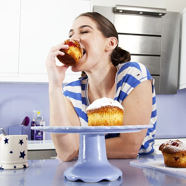 Naine kodus köögis muffinit süües, laual on veel magusat küpsetist ja tass